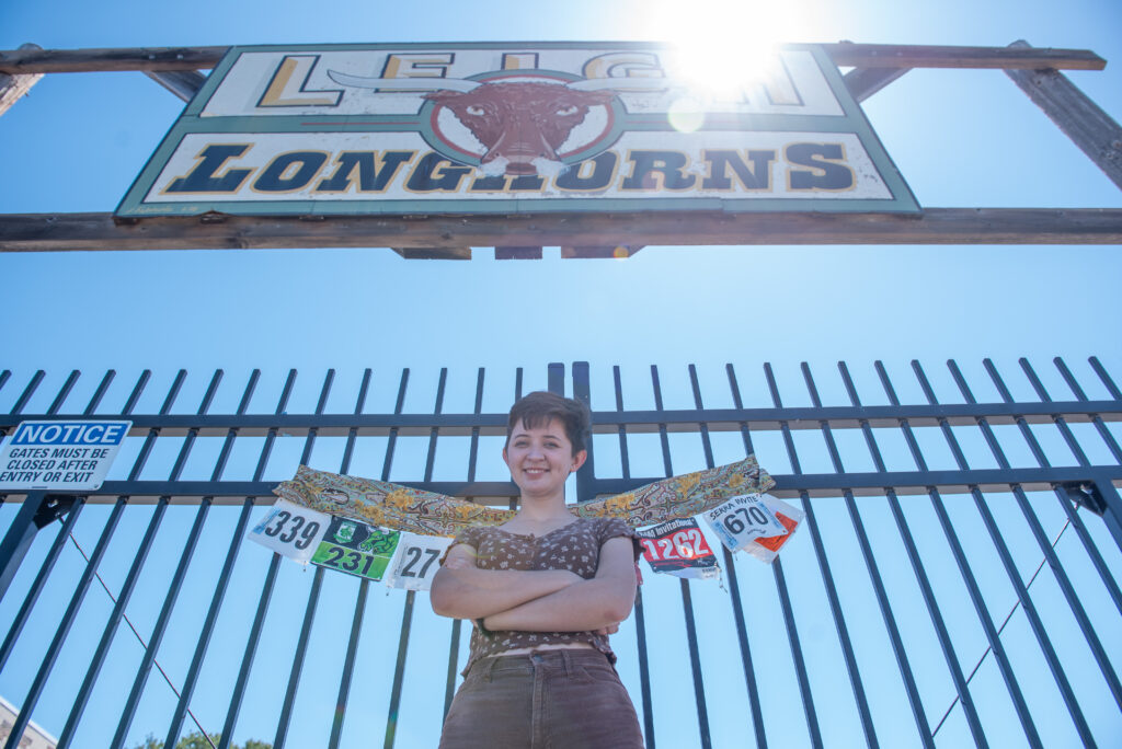 Teen female arm across her chest posing in front of school gates with cross country bib numbers displayed on the gate. 