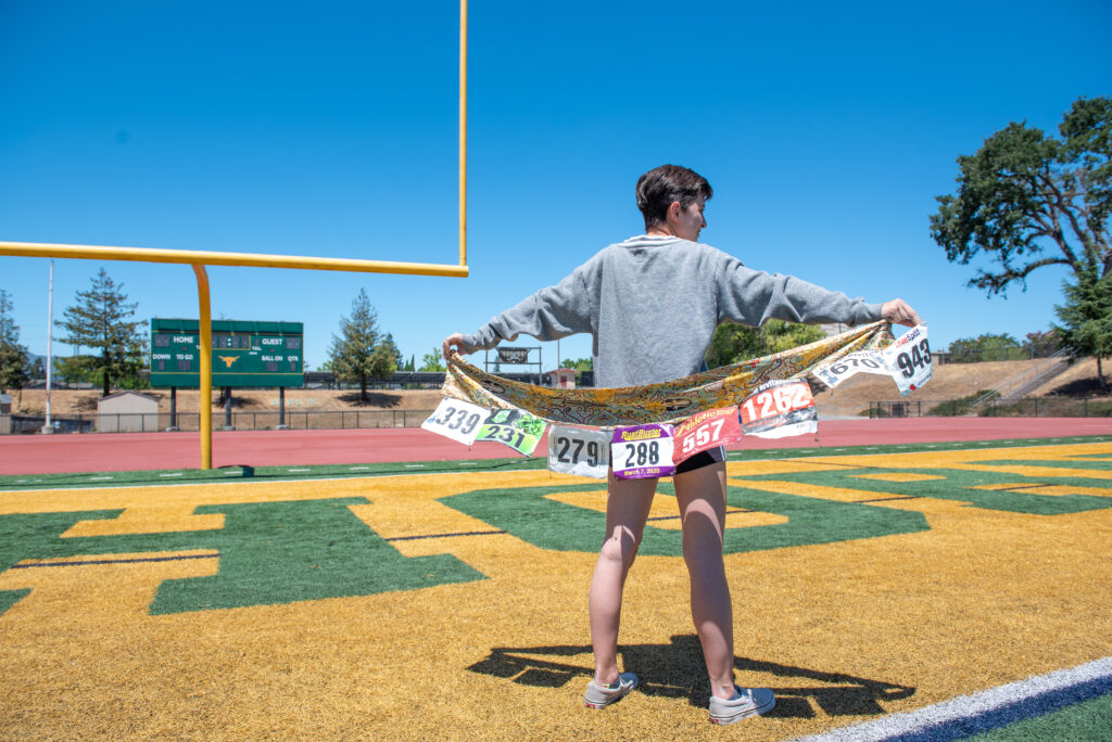 Teen female girl back is turned away while holding banner of cross country bib numbers behind her back spread across both arms.