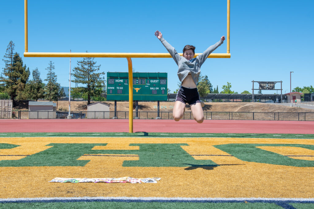 Teen female girl jumps in the air with two arm high on high school football field end zone.