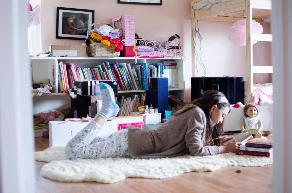 Female tween girl reads books in her room lying on her stomach, as captured looking through the doorway.